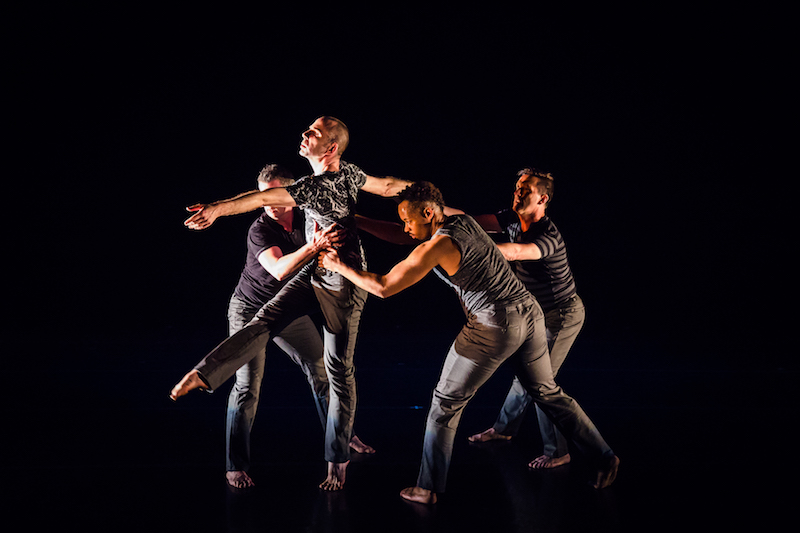 Three dancers hold the torso of a fourth dancer. The focal dancer balances on one leg with their other leg outstretched. 